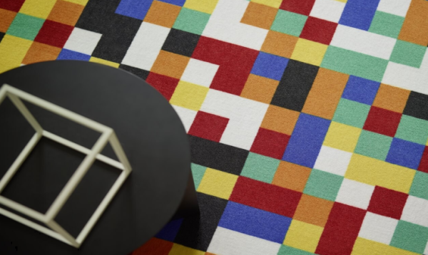 „Bunter Vorwerk Teppich mit pixelartähnlichem Quadratmuster in Rot, Gelb, Blau, Grün, Orange, Schwarz und Weiß; im Vordergrund schwarzer Tisch mit weißem Drahtwürfel auf grauem Boden.“